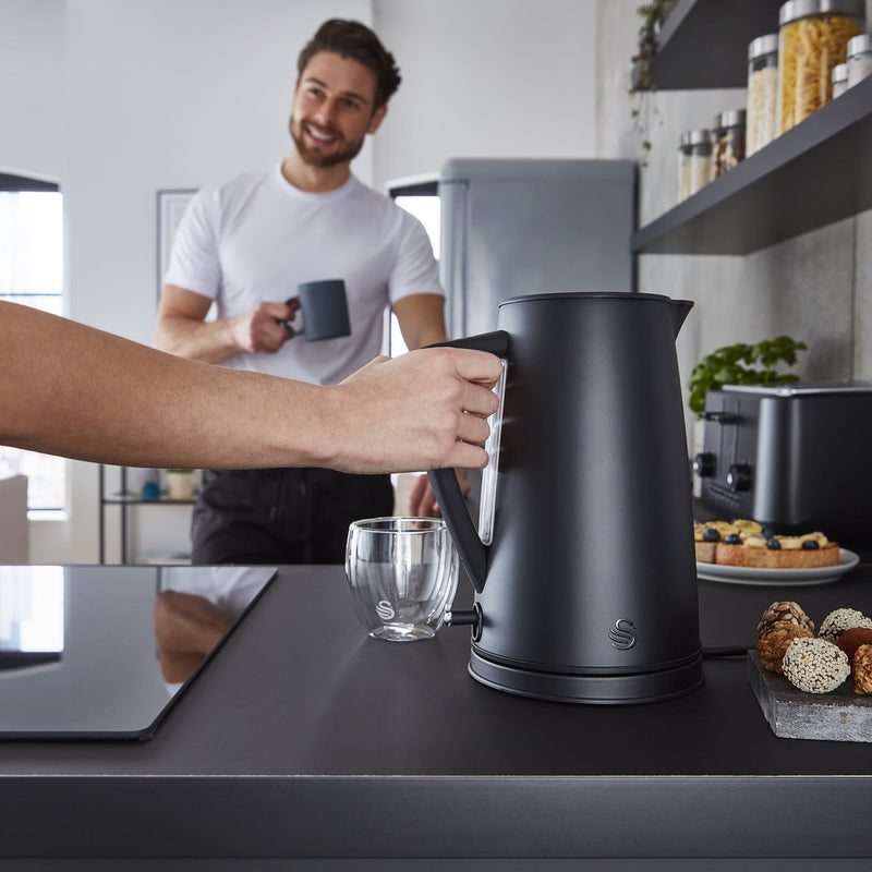 Photograph of Swan's 1.7 Litre Stealth Kettle next to assorted food and a man behind holding a coffee mug in a modern kitchen
