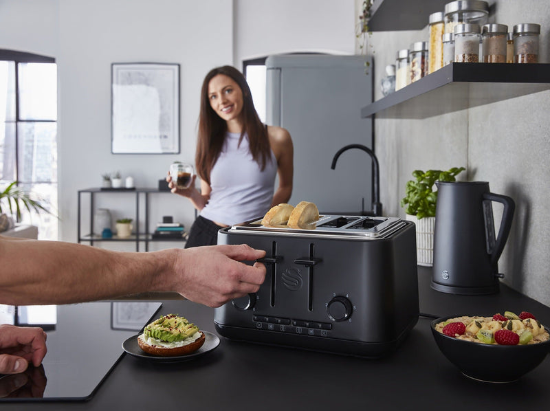 Distanced shot of a woman holding a glass of coffee in a modern kitchen with a Swan Stealth 4 slice toaster being used by a man, next to a bowl of cut up fruit and a sliced bagel with smashed avocado.