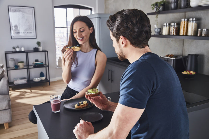 Photograph of a woman holding toast with blueberries and banana toppings and a man holding a bagel with cream cheese and smashed avocado toppings in a modern kitchen with Swan's Stealth 4 Slice Toaster in the background 