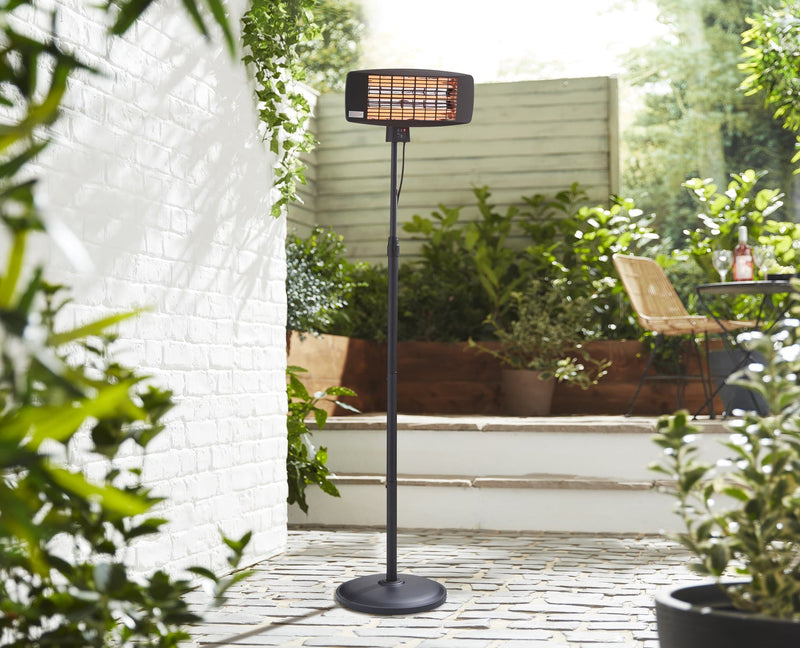 Full length shot of the Swan Stand Patio Heater on a cobblestone patio, with green plants in the distance.