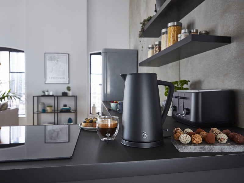 Photograph of a Swan 1.7 Litre Stealth Kettle next to a glass of coffee and assorted foods in a modern kitchen