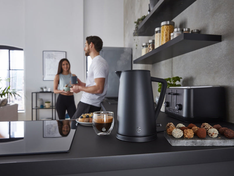 Photograph of a Swan 1.7 Litre Stealth Kettle next to a glass of coffee and assorted foods with a man and woman drinking and eating behind in a modern kitchen