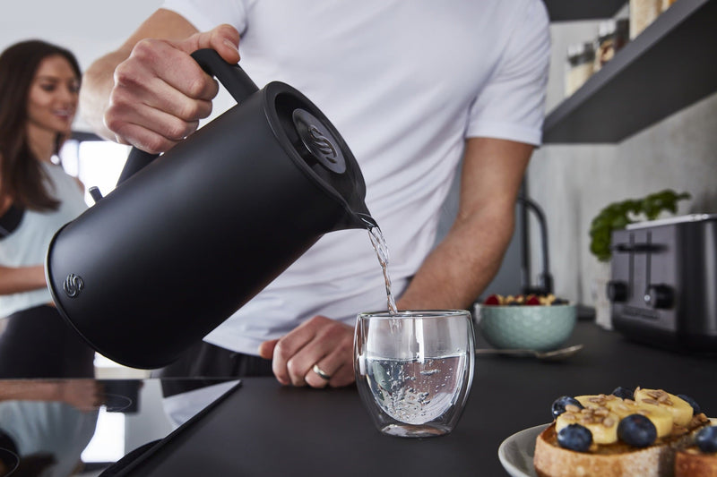 Photograph of man pouring boiling water from a Swan 1.7 Litre Stealth Kettle into a small transparent glass next to assorted foods in a modern kitchen