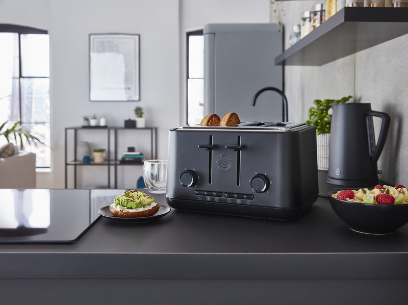 Distanced shot of Swan's Stealth 4 slice toaster in a modern kitchen, next to a bowl of cut up fruit and a sliced bagel with smashed avocado.