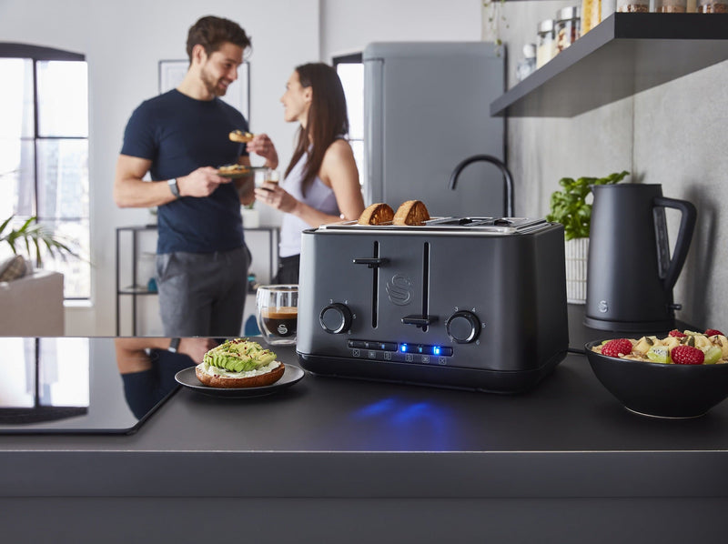 Distanced shot of a man and a woman eating and drinking in a modern kitchen with a Swan Stealth 4 slice toaster, next to a bowl of cut up fruit and a sliced bagel with smashed avocado.