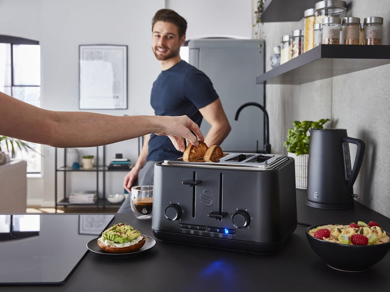 Distanced shot of a man in a modern kitchen with a Swan Stealth 4 slice toaster being used by a woman, next to a bowl of cut up fruit and a sliced bagel with smashed avocado.