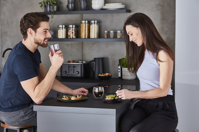 Photograph of a man and woman sitting next to a modern kitchen work top with assorted foods on with Swan's Stealth 4 Slice Toaster in the background