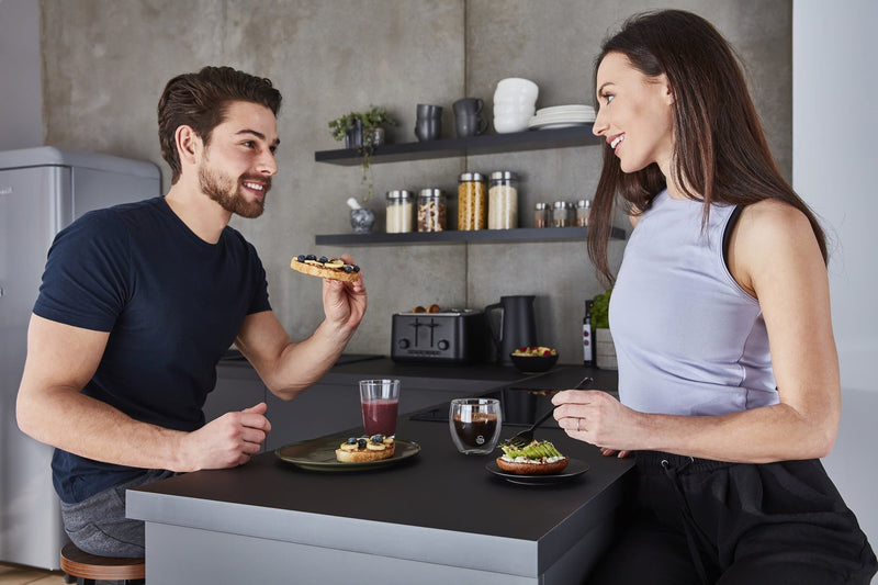 Photograph of a man holding toast with blueberries and banana toppings and a woman holding a fork above a bagel with cream cheese and smashed avocado toppings in a modern kitchen with Swan's Stealth 4 Slice Toaster in the background